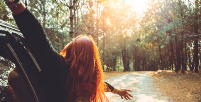 A girl hanging out of a car window on a forested road.