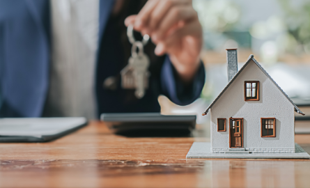 A miniature house in the forefront with a person holding keys in the background.