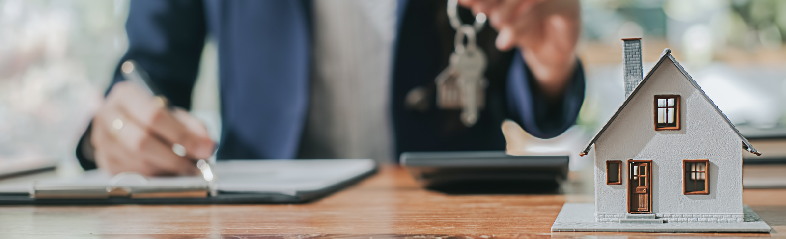 Miniature house in forefront with a person holding a set of house keys writing on a notepad.