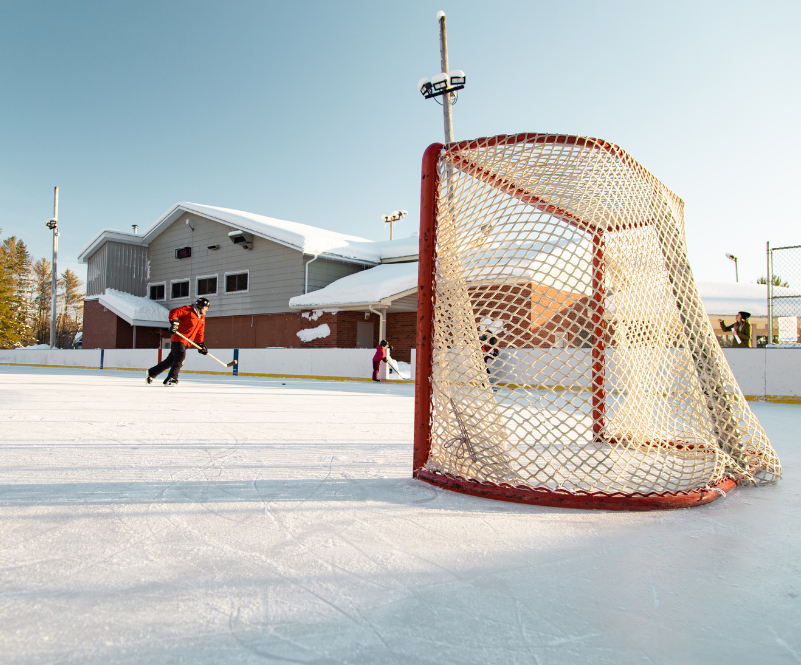 People playing hockey on an outdoor hockey rink with the recreation centre in the background.